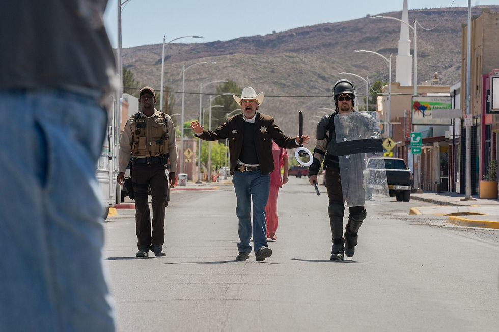Joaqin Phoenix and his men walk down a sunny street in a small town. One wears a cowboy hat, another in tactical gear, and the third in riot gear.