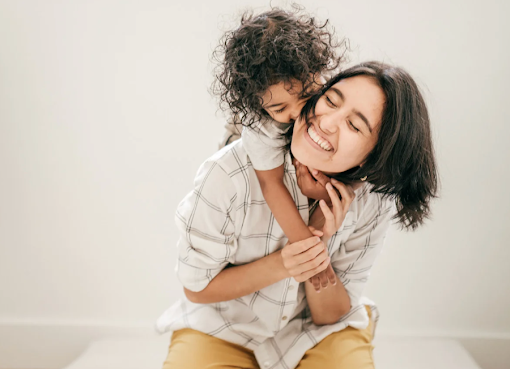 Mother smiling as her child hugs her from behind, showing the warmth and closeness of family life alongside work responsibilities.