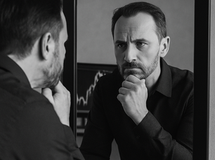 A black-and-white photograph of a middle-aged man with a thoughtful expression, gazing at his reflection in a mirror. He rests his chin on his hand, appearing deep in contemplation. A blurred financial chart is visible in the background, hinting at trading or market analysis.