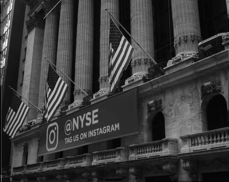 Black-and-white photograph of the New York Stock Exchange building with large American flags and a banner reading “@NYSE TAG US ON INSTAGRAM.