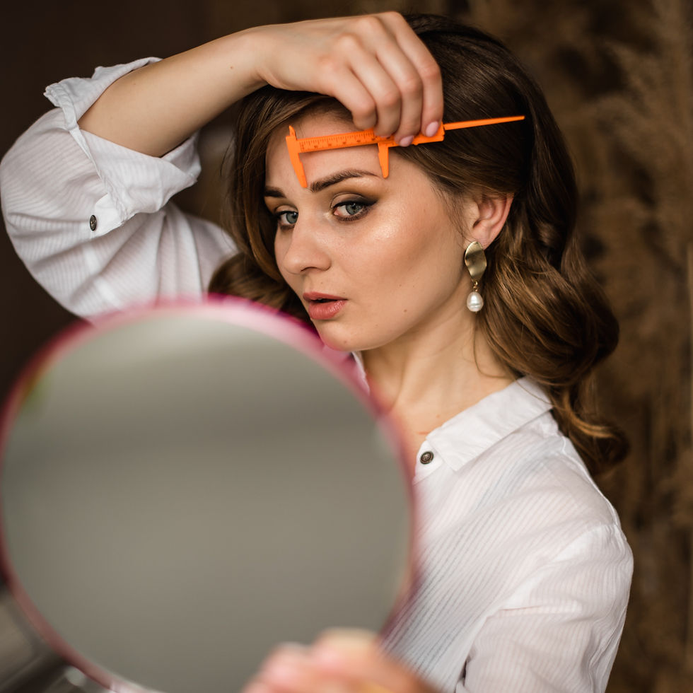 Woman in white shirt using an orange eyebrow ruler, looks in a mirror. Focused expression, brown background, elegant earrings.