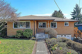 Here you can see a single story brick home, with a drive-under garage to the right. The brick is a light tan and you can see 4 windows on the front of the house. A flower garden is to the right of the brick-lined path leading to the front door. Poking out of the right side of the home you can see a wooden deck. 