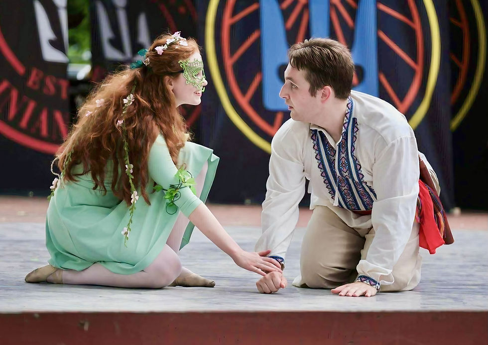 Two performers in colorful costumes kneel on stage, one in a green dress and mask, the other in embroidered attire. Focused expressions.