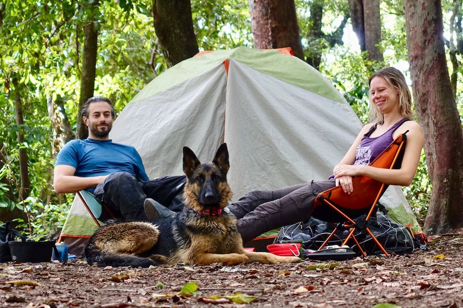 A couple and dog sit beside tent at campsite in the woods