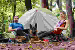 A couple and dog sit beside tent at campsite in the woods