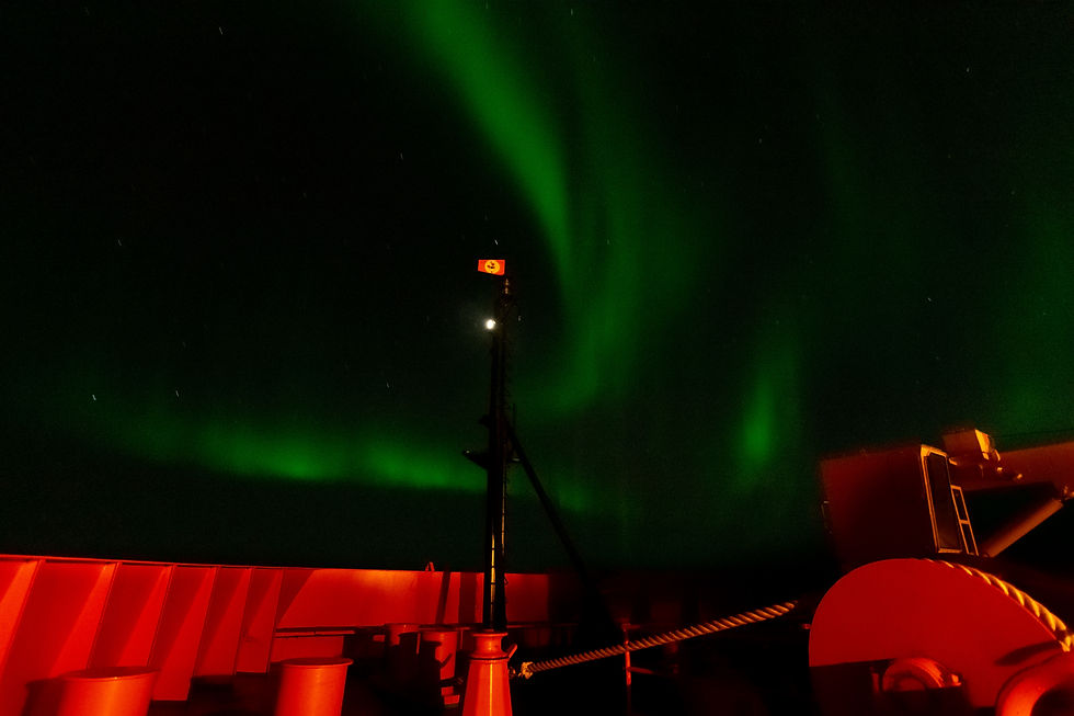 Green bands of light arch across a night sky in antarctica. Flag at front of US reaearch vessel in center of photo. Red walls of boat at bottom of picture. Photo by: Chandler Olson