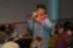 Musician Klassik performs an improvised song on a mic amongst audience members seated at tables.