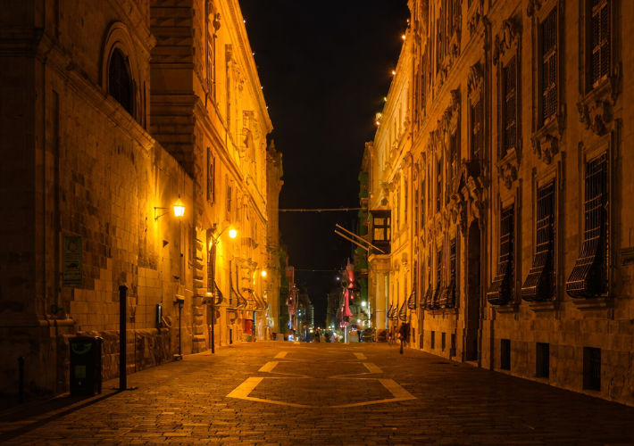 Narrow historic Maltese street at night illuminated by warm street lamps, with traditional stone buildings lining both sides.