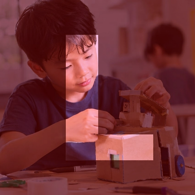 Digital photography of a young boy intently building a cardboard model. The scene suggests a creative and educational activity.
