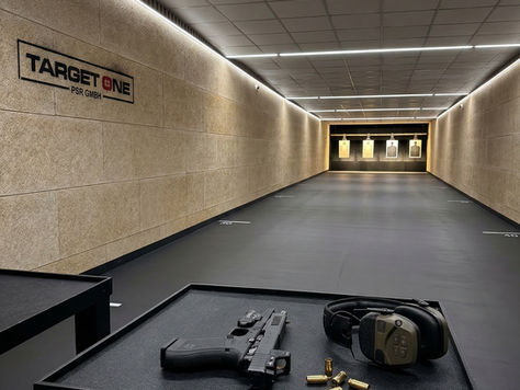 Professionelle Aufnahme eines modernen Indoor-Schießstandes der Marke Target One PSR. Im Vordergrund liegen eine schwarze taktische Pistole mit Optik, Kapselgehörschutz und leere Patronenhülsen auf einem schwarzen Tisch. Der Blick führt entlang einer hell erleuchteten Schießbahn mit sandfarbenen Akustikwänden hin zu beleuchteten Zielscheiben im Hintergrund.