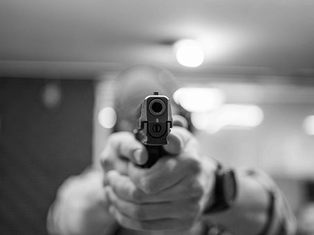 Eine Person hält eine Pistole mit beiden Händen direkt auf die Kamera gerichtet. Im Vordergrund ist der Lauf der Waffe scharf zu sehen, während Hintergrund und Gesicht unscharf bleiben. Dramatische Perspektive mit Fokus auf den Mündungsbereich.