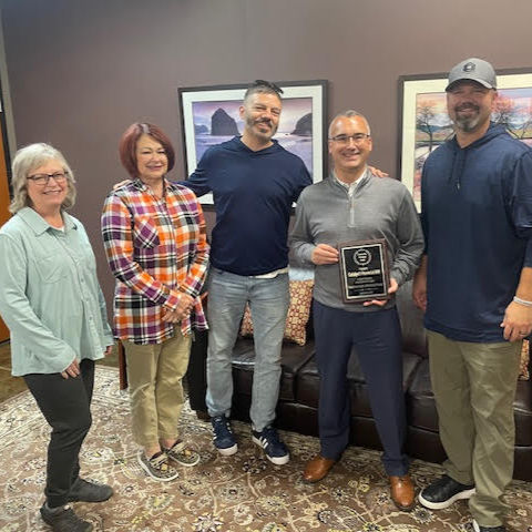 Five adults stand together in an office setting, smiling for a photo. The man in the center holds a black thank-you plaque presented to the Catalyst Financial NW team in recognition of their support for Transitional Youth and partner nonprofits. Framed landscape photos hang on the wall behind them, and the group stands on a patterned rug in front of a brown leather couch.