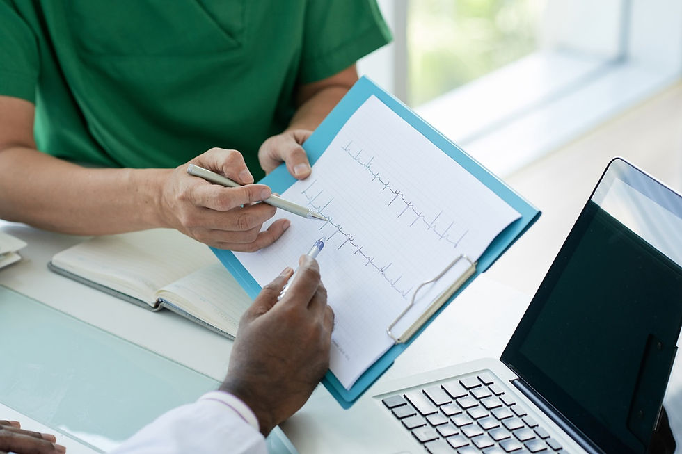 Two people discuss an EKG chart on a clipboard in a bright office. One wears green scrubs, pointing with a pen near a laptop.