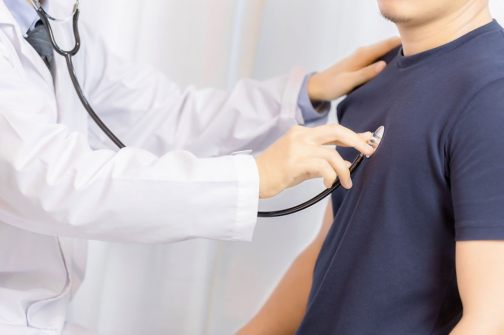Doctor in white coat using a stethoscope to check a patient's heartbeat in a clinical setting. The patient wears a navy shirt.