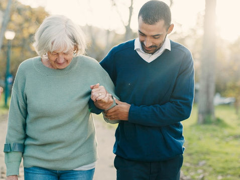 Elderly woman and man walk arm in arm in a sunlit park, both looking down with a calm, caring mood.
