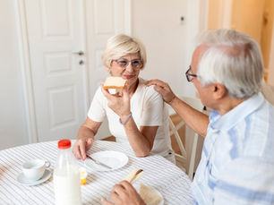 Elderly couple enjoying breakfast at a table with striped cloth. Woman holds bread, man gently touches her shoulder, creating a warm atmosphere.