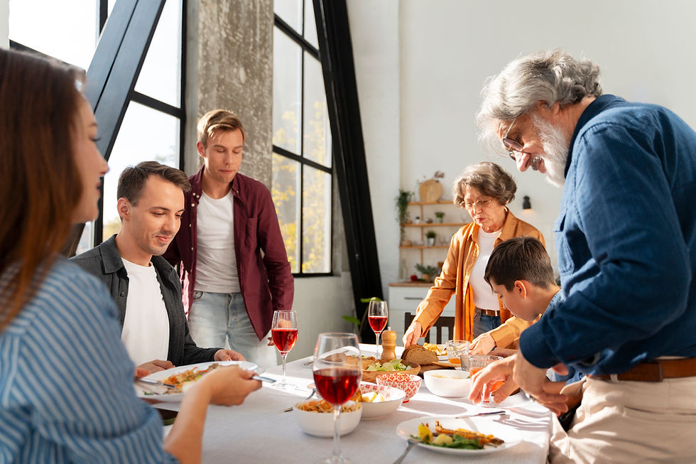 Family dining together at a table with plates of food and glasses of wine. Sunlit room with large windows and shelves in the background.