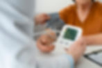 Doctor checks a patient's blood pressure with a monitor. The patient wears an orange shirt. The setting appears to be a clinic.