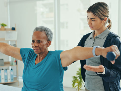 Elderly woman in a blue shirt lifts dumbbells with a physical therapist guidance in a bright room, looking focused and determined.