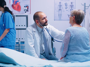 Doctor with stethoscope listens to patient in blue top in a clinic. Nurse in scrubs on computer. Medical charts on walls.