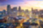 City skyline at sunset with tall buildings, glowing windows, and vibrant sky hues of orange and purple. Streetlights illuminate below.