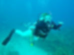 Scuba diver in black wetsuit making a hand gesture underwater, surrounded by blue hues and coral reef, conveying excitement.
