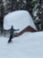 Snowboarder poses playfully on snowy slope near a cabin with a massive snow mound. Snow-covered trees in the background.