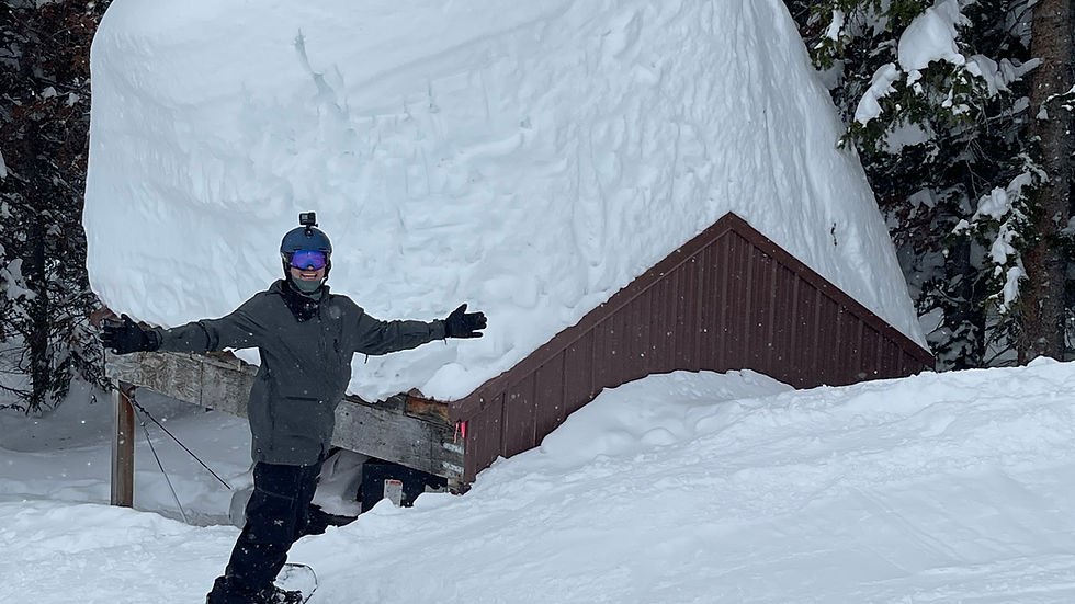 Snowboarder poses playfully on snowy slope near a cabin with a massive snow mound. Snow-covered trees in the background.