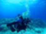 Scuba diver in blue wetsuit swimming underwater with coral reef backdrop. Clear blue water, bubbles rising, and peaceful atmosphere.