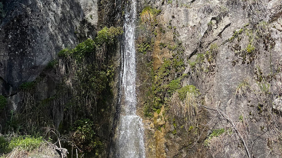 Waterfall cascading down a rocky cliff surrounded by greenery. Clear blue sky above, creating a serene, natural setting.
