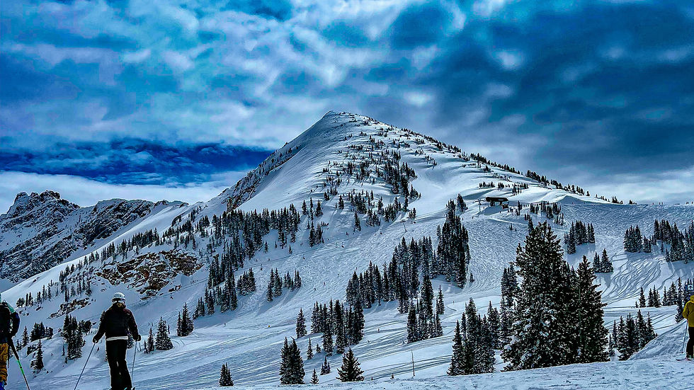 Skiers on snowy slope with pine trees, under a vivid blue, cloudy sky. Sunlight peeks through, creating a serene winter scene.