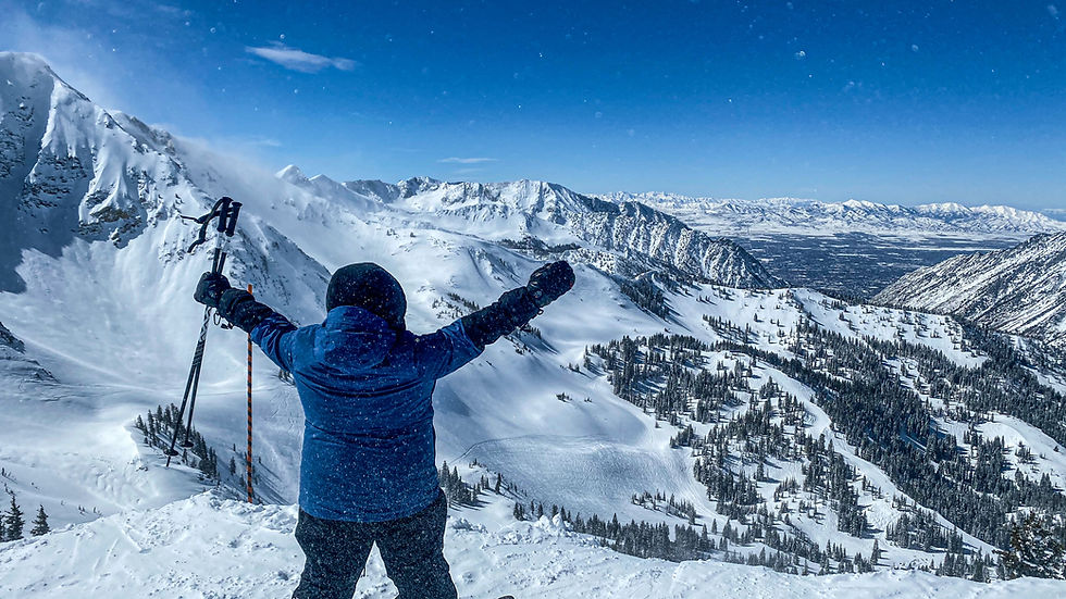 Skier in blue jacket raises arms triumphantly atop a snowy mountain with ski poles; clear blue sky and expansive snowy landscape.