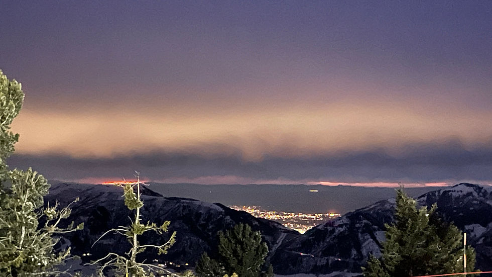 Snowy mountain landscape at dusk with distant city lights glowing under a purple-orange sky. Foreground trees add depth. Calm mood.