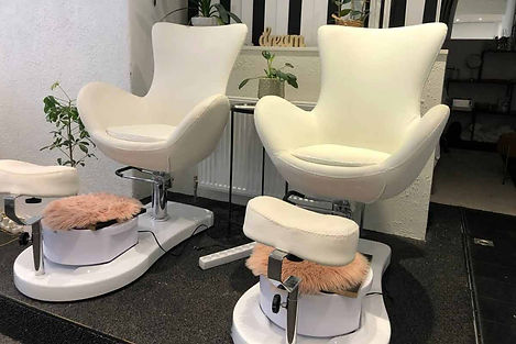 To hvide pedicurestole med fodstøtter i et salonmiljø. Stolene har et moderne design med buede armlæn og er udstyret med fod