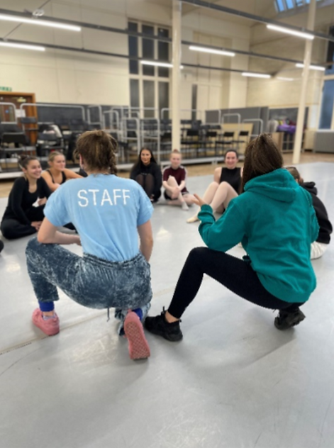 In the background, a higher dance class sat in a semi circle. In the foreground, two staff members crouching and speaking with their backs to the camera. One with staff on the back of their t-shirt.