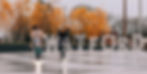 Two women ride scooters in front of a WATFORD sign with autumn trees.