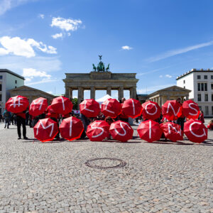 People holding red umbrellas with "#MILLIONS MISSING" in white, standing in a sunny plaza near the Brandenburg Gate.