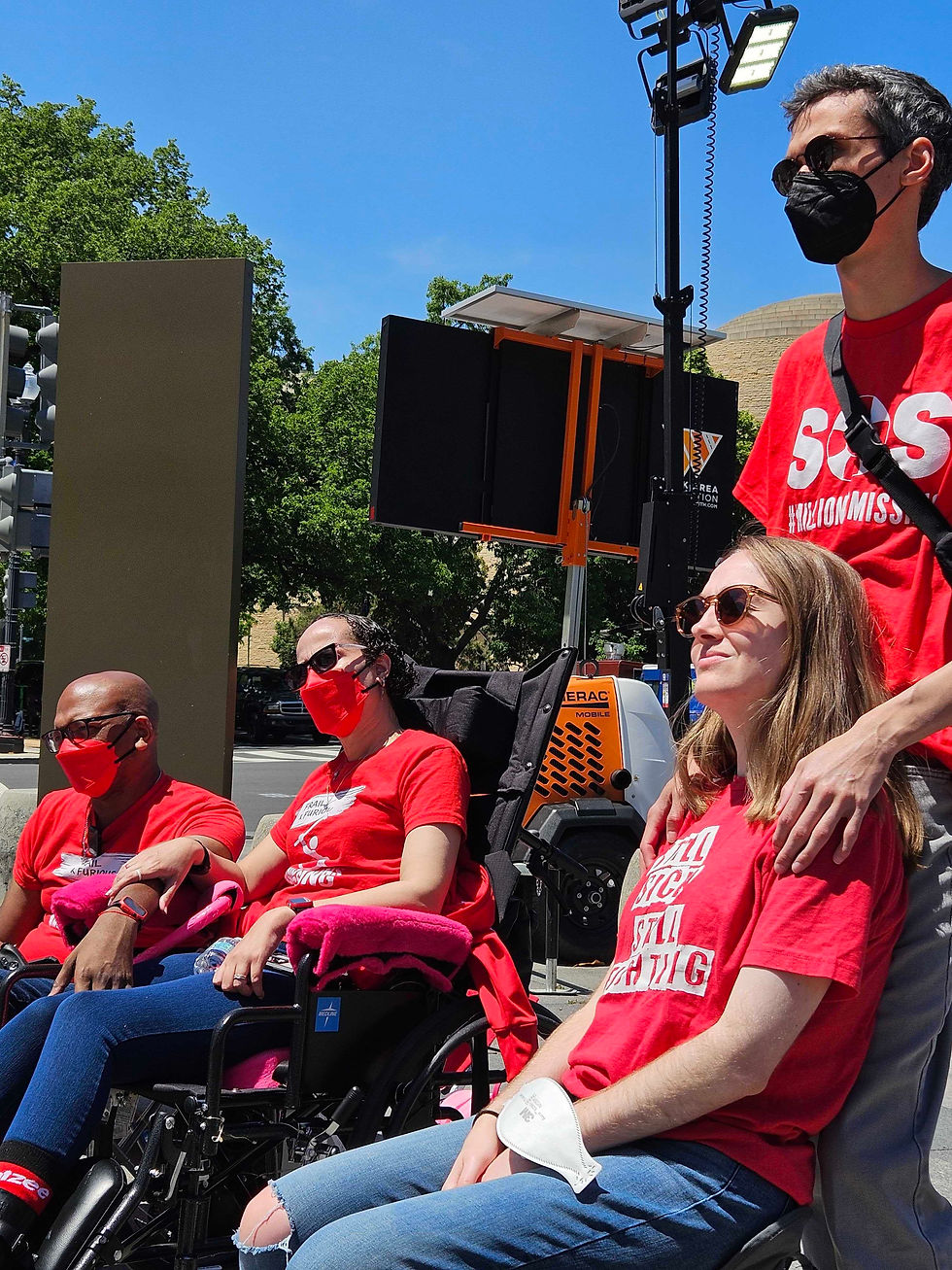 Four people in red shirts, some in wheelchairs, participate in an outdoor event. A person stands with hand on another's shoulder. Sky is clear.