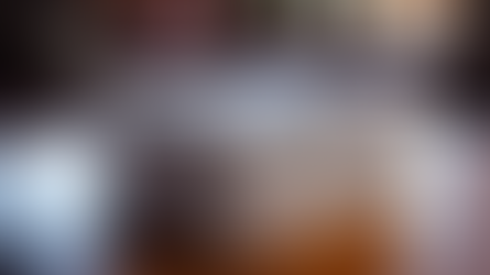 Stack of newspapers on a wooden table in a cozy living room with warm lighting. A sofa and plant are blurred in the background.