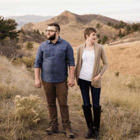 A couple holding hands walks on a grassy trail. Hills and trees in the background. They look content. The man wears a denim shirt.