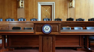 Wooden podium with "United States Senate" seal in an empty chamber, surrounded by leather chairs. Warm lighting, formal atmosphere.
