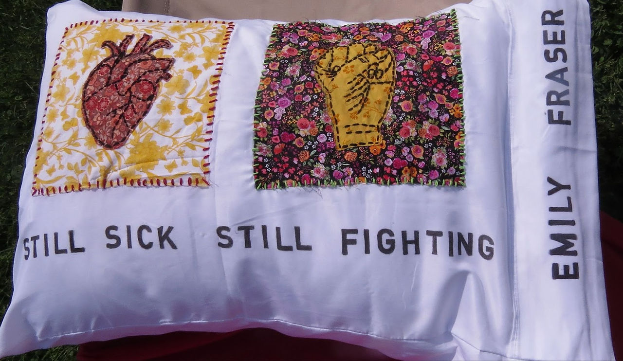 White fabric with embroidered patches: a heart on yellow floral, a fist on black floral. Text: "STILL SICK STILL FIGHTING" and "EMILY FRASER".