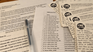 Letters, printed lists of addresses, and handwritten postcards with #MEAction stickers are spread on a table. A pen is also visible.