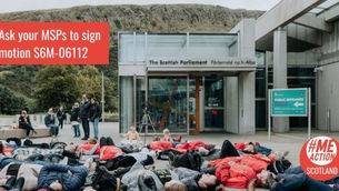 People lie down in red clothing in front of the Scottish Parliament. A sign reads "Ask your MSPs to sign motion S6M-06112."