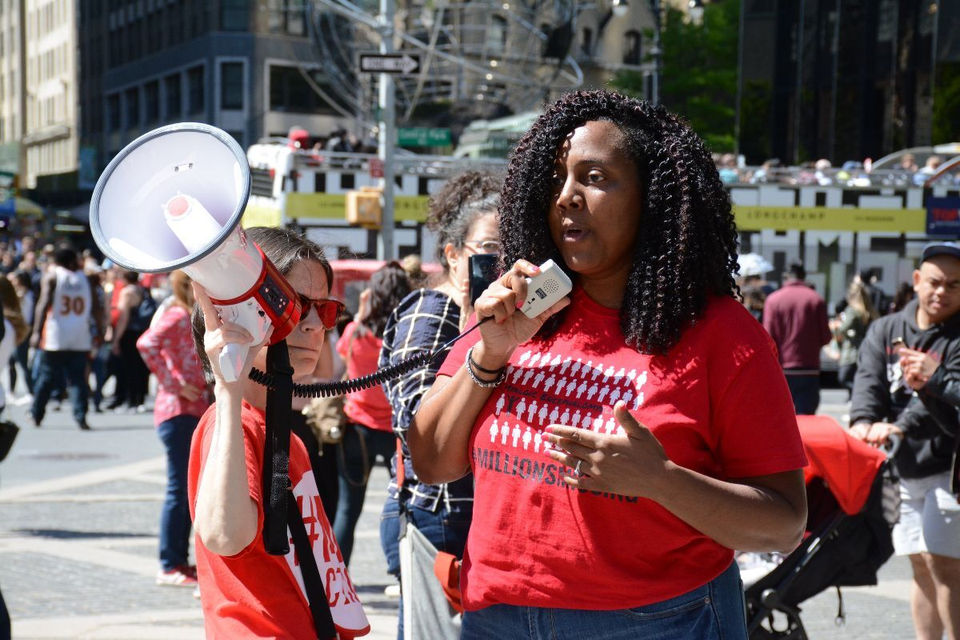 A woman in a red shirt passionately speaks into a megaphone, conveying a message to a gathered crowd.