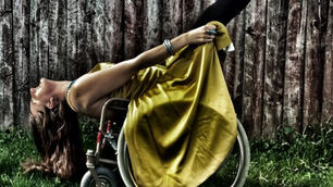 Woman in a yellow dress gracefully leans back in a wheelchair on grass, against a wooden fence background, exuding elegance and confidence.