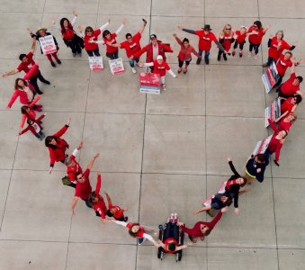 People in red shirts form a heart shape on a concrete surface, holding signs. The mood is cheerful and supportive.