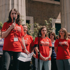 A woman speaks into a microphone at a #MillionsMissing demonstration. A group of people wearing red #MillionsMissing shirts stand behind her.
