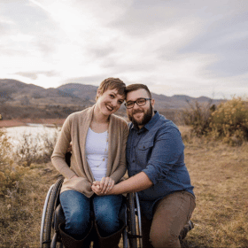 A couple smiles in a natural setting, with mountains and a lake in the background. The woman is in a wheelchair, and the mood is joyful.
