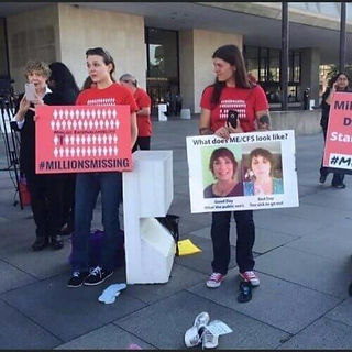 People in red shirts hold signs about ME/CFS awareness outside a building. Signs feature images and hashtags like #MILLIONSMISSING.